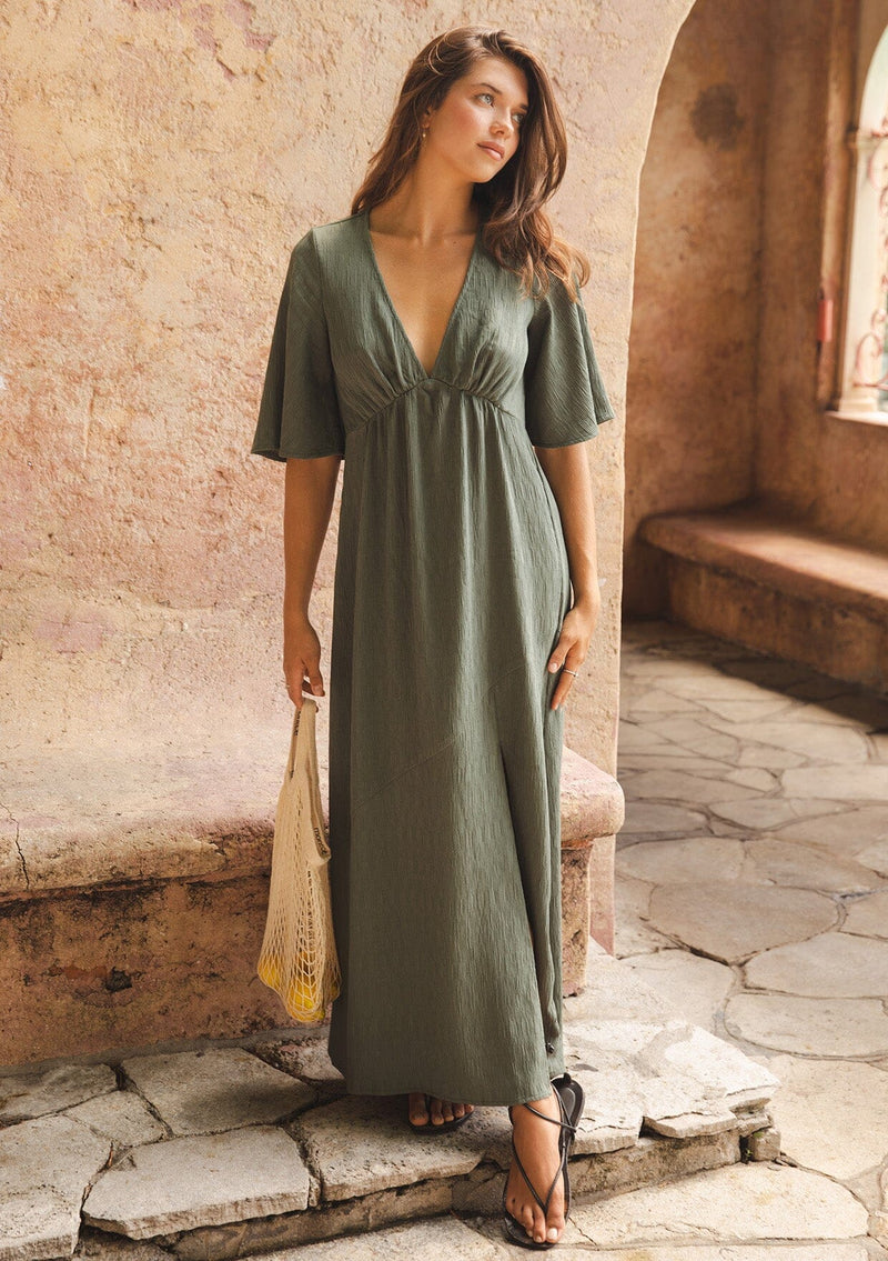 [Color: Olive] A brunette model wearing a textured maxi dress in a green autumn hue. With a deep v neckline, empire waistline, flowy half sleeves, and a side slit. Paired with casual sandals for any daytime outing.