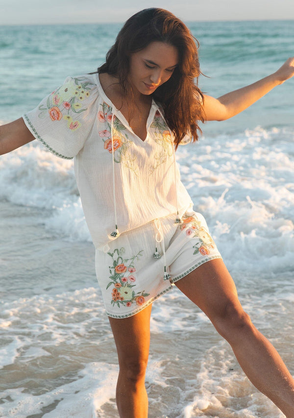 [Color: Cream/Coral] A brunette model wearing a cotton cream top with contrasting floral embroidered details. This relaxed cotton top features short sleeves and a split v-neckline with ties. Perfect for lounging or vacations. 