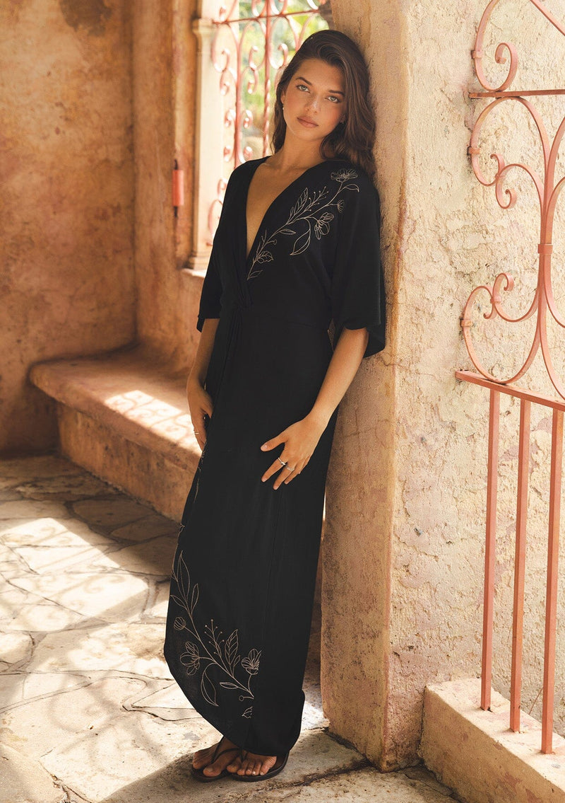 [Color: Black/Natural] A brunette model wearing a black front knot maxi dress with a contrast embroidery, Featuring a deep v neckline, kimono sleeves, front slit, and an asymmetrical hem. A bohemian style maxi dress with a relaxed fit.  