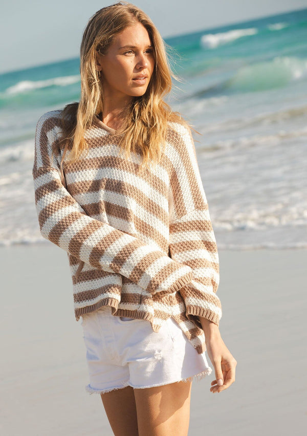 [Color: Natural/Taupe] A front facing image of a blonde model wearing a natural and taupe striped knit hoodie at the beach. With long sleeves, a dropped shoulder, a v neckline, and a front kangaroo pocket. 