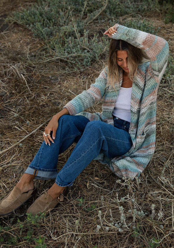 [Color: Mint/Natural] A sitting image with a blonde model wearing a cozy bohemian space dye chunky knit cardigan in a green mint multi-color stripe pattern. This plush cardigan features long sleeves, a cozy hood, essential pockets, and an open front. The long length and thick knit make it the perfect sweater for the fall season.