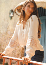 [Color: Natural/Taupe] A brunette model wearing an off white bohemian blouse with embroidered details. Featuring a split v neckline, self covered button front, and long sleeves with smocked cuffs. Paired with light wash denim for the perfect fall look.