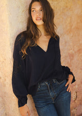 [Color: Navy] A lifestyle image of a brunette model wearing a bohemian navy blue blouse. With a split v neckline, long sleeves, ruffled elastic wrist cuffs, and embroidered lace details. A fall blouse styled with denim and boots. 