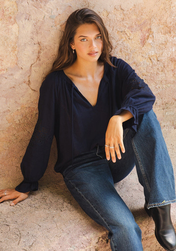 [Color: Navy] A lifestyle image of a brunette model wearing a bohemian navy blue blouse. With a split v neckline, long sleeves, ruffled elastic wrist cuffs, and embroidered lace details. A fall blouse styled with denim and boots. 