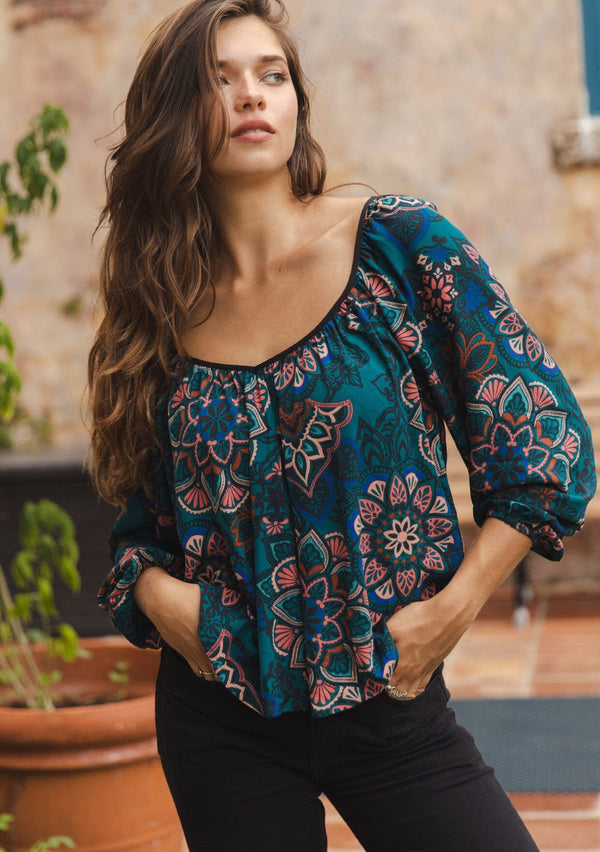 [Color: Teal/Rust] A brunette model wearing an ultra-flowy boho blouse in a teal floral print with contrast piping. Featuring a wide v neckline, long sleeves, and an open back with tie detail.