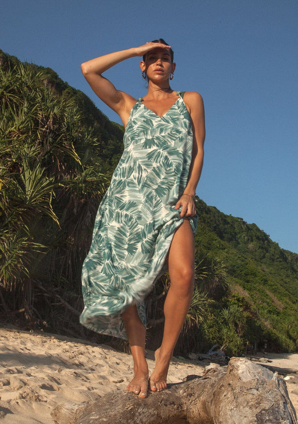 [Color: Natural/Green] A brunette  model on a tropical island wearing a sleeveless summer maxi dress in a tropical green palm leaf print. With a v neckline, spaghetti straps with bead accents, a strappy back with adjustable tie, and a flowy silhouette. 