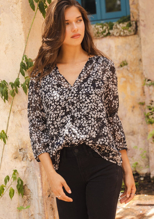 [Color: Black/Grey] A brunette woman wearing an ultra flowy chiffon blouse with metallic clip dot details. A black floral blouse with a split v neckline, long sleeves, and elastic cuffs. Paired with denim jeans for a relaxed fall look, perfect for dinner and classy evening parties.