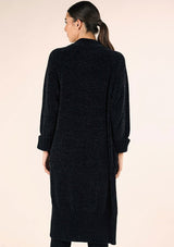 [Color: Black] A back facing image of a brunette model wearing a soft black chenille knit duster cardigan. With long sleeves, an open front, and side pockets. 