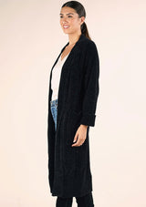 [Color: Black] A side facing image of a brunette model wearing a soft black chenille knit duster cardigan. With long sleeves, an open front, and side pockets. 