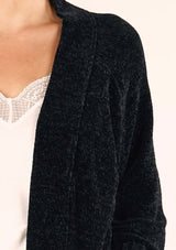 [Color: Black] A close up front facing image of a brunette model wearing a soft black chenille knit duster cardigan. With long sleeves, an open front, and side pockets. 