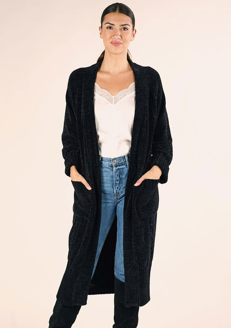 [Color: Black] A front facing image of a brunette model wearing a soft black chenille knit duster cardigan. With long sleeves, an open front, and side pockets. 