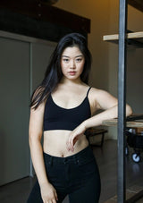 [Color: Black] A brunette model wearing a fitted crop lingerie top in dark black. A casual yet stylish undergarment perfect for styling under dresses or wearing on it's own as a sports bra. A comfortable stretchy ribbed knit top with a low back and spaghetti straps.