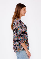 [Color: Black/Light Blue] A brunette woman wearing a multi color floral print top with long sleeves and a wide v neckline. A casual top tucked in styled with denim pants. A summer to fall style for casual outings. 