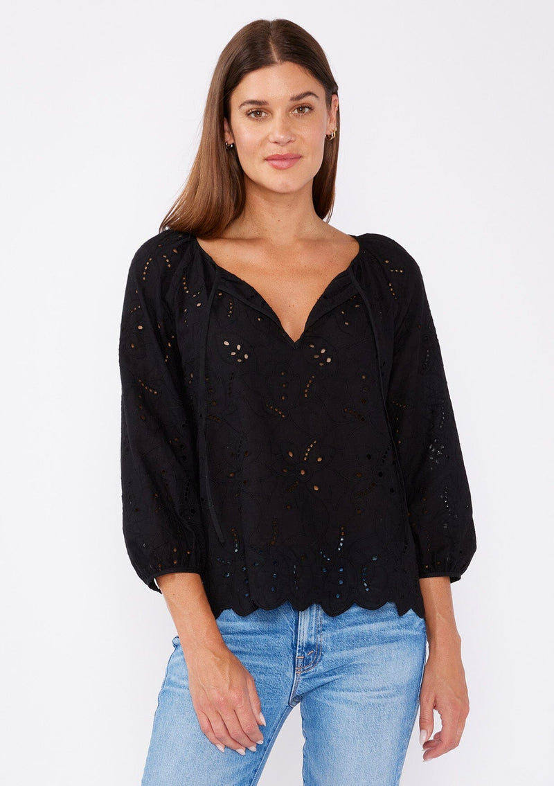[Color: Black] A brunette woman wearing an embroidered eyelet top with a split v neckline with ties, scalloped hem, and three quarter sleeve with button cuff. A see through top with no lining. 