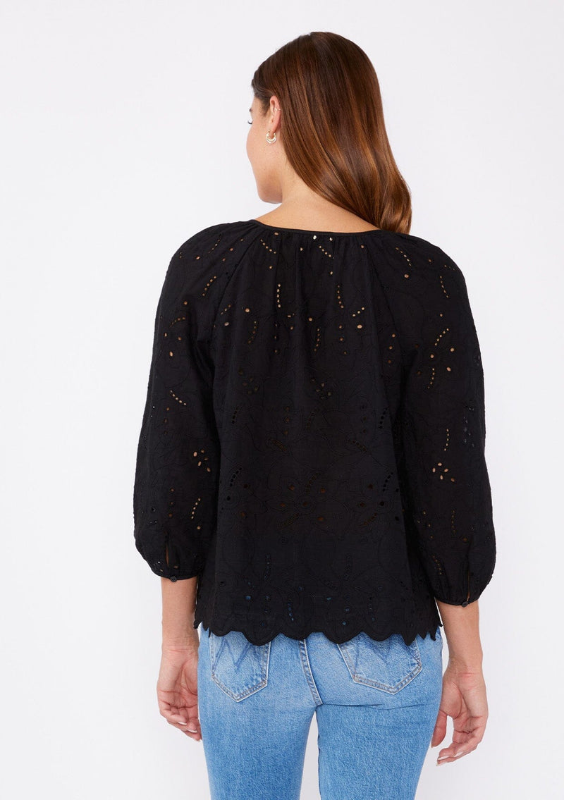 [Color: Black] A brunette woman wearing an embroidered eyelet top with a split v neckline with ties, scalloped hem, and three quarter sleeve with button cuff. A see through top with no lining.