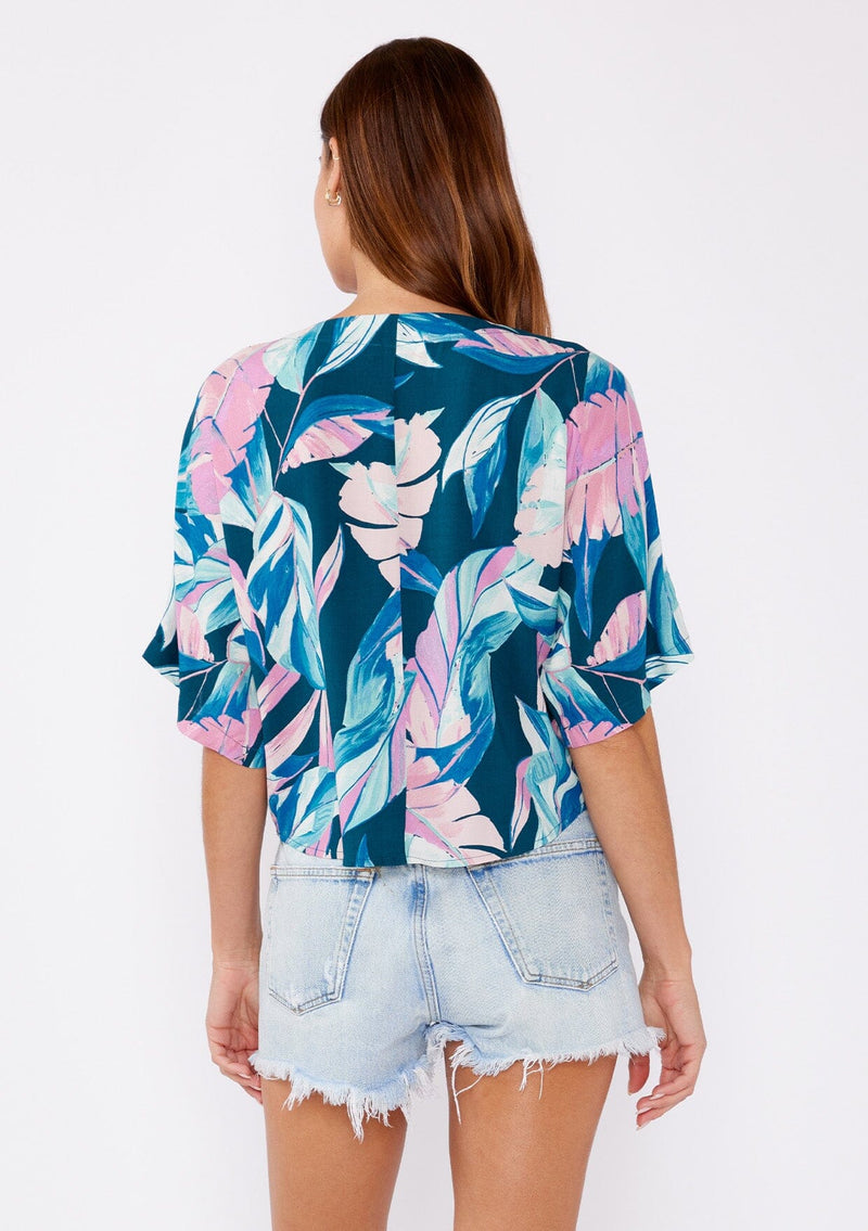 [Color: Teal/Dusty Orchid] A brunette model wearing a tropical leaf print top. This twist front top features a button up front, short dolman sleeves, and a v neckline. A casual top paired with denim shorts.