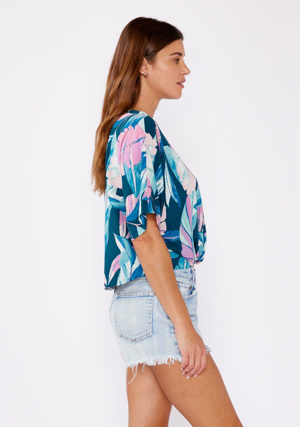 [Color: Teal/Dusty Orchid] A brunette model wearing a tropical leaf print top. This twist front top features a button up front, short dolman sleeves, and a v neckline. A casual top paired with denim shorts.