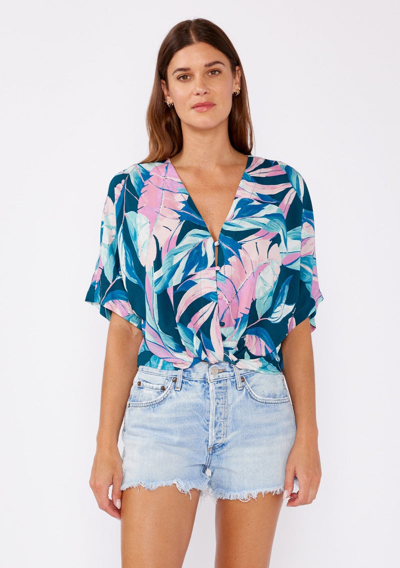 [Color: Teal/Dusty Orchid] A brunette model wearing a tropical leaf print top. This twist front top features a button up front, short dolman sleeves, and a v neckline. A casual top paired with denim shorts.