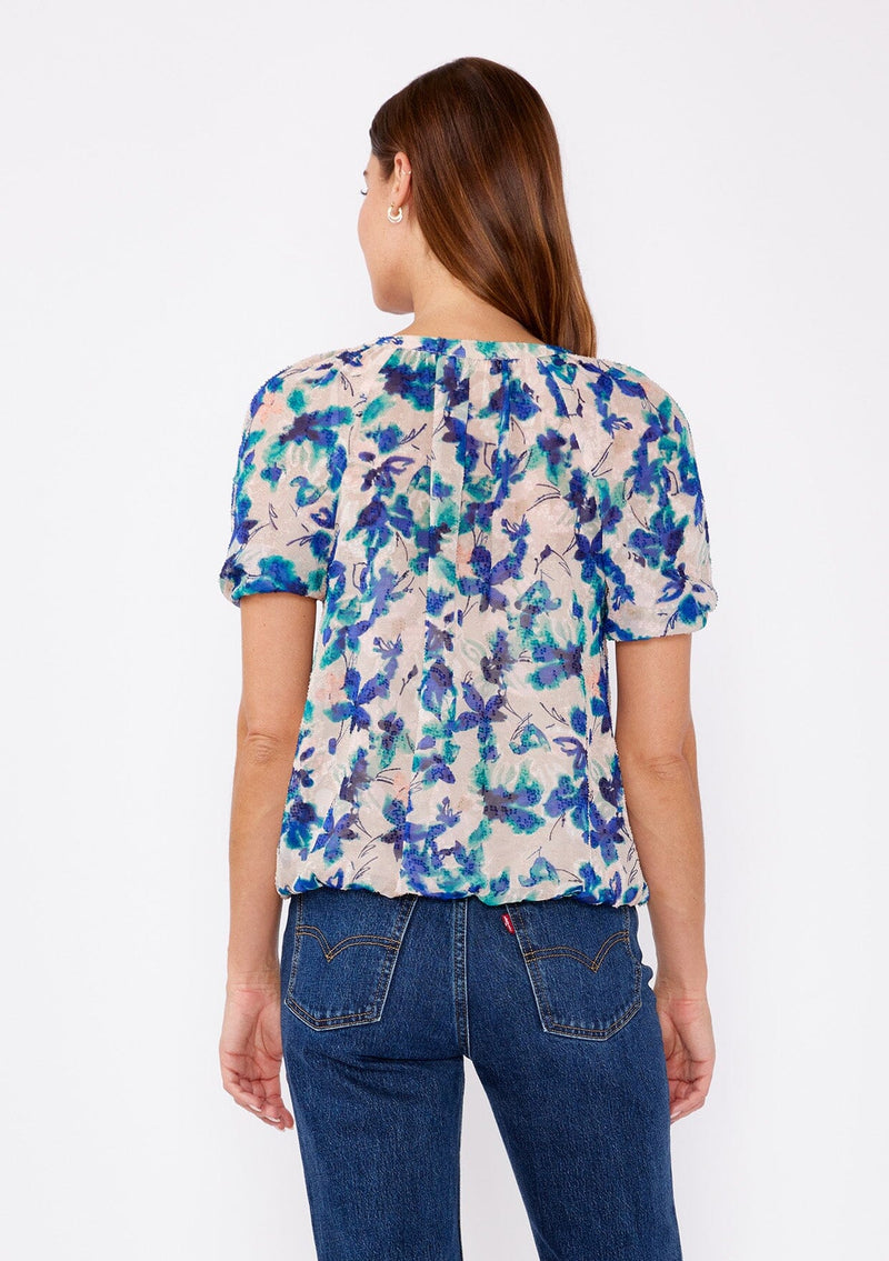 [Color: Natural/Teal] A brunette woman wearing a blue and natural floral chiffon blouse with puff sleeves, decorative button front, and a elastic hem. A summer casual top to wear with denim jeans to dinner or special occasions.