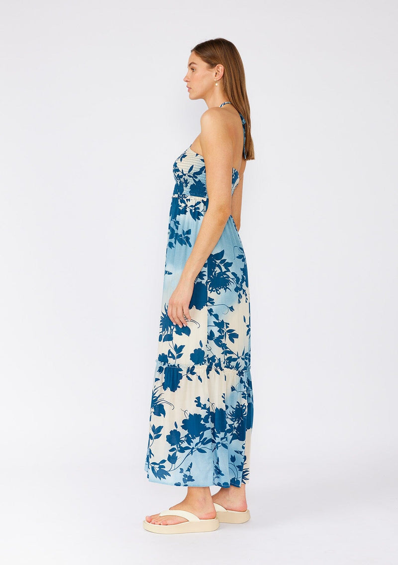 [Color: Navy/Dusty Blue] A side facing image of a brunette model wearing a dreamy bohemian halter maxi dress in a blue floral print. With a smocked elastic slim fit bodice, a long ruffles trimmed tiered skirt, and an adjustable halter tie neckline. 