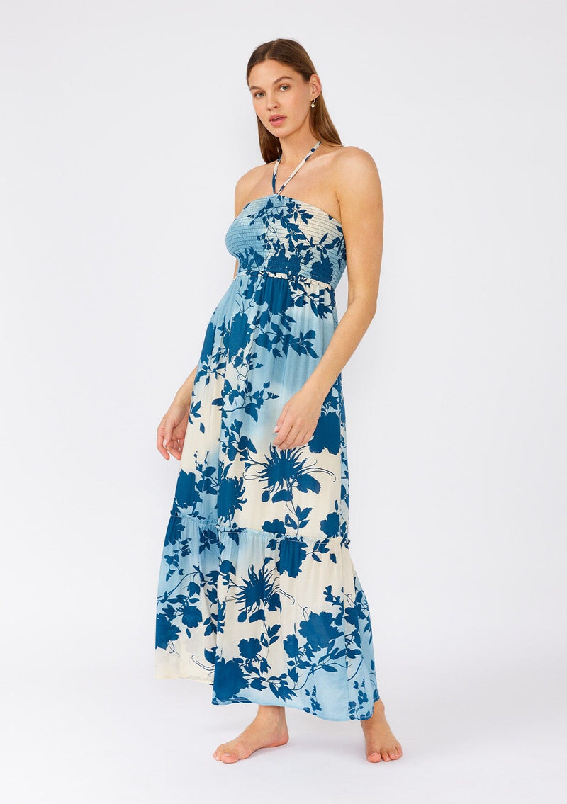 [Color: Navy/Dusty Blue] A full body front facing image of a brunette model wearing a dreamy bohemian halter maxi dress in a blue floral print. With a smocked elastic slim fit bodice, a long ruffles trimmed tiered skirt, and an adjustable halter tie neckline. 