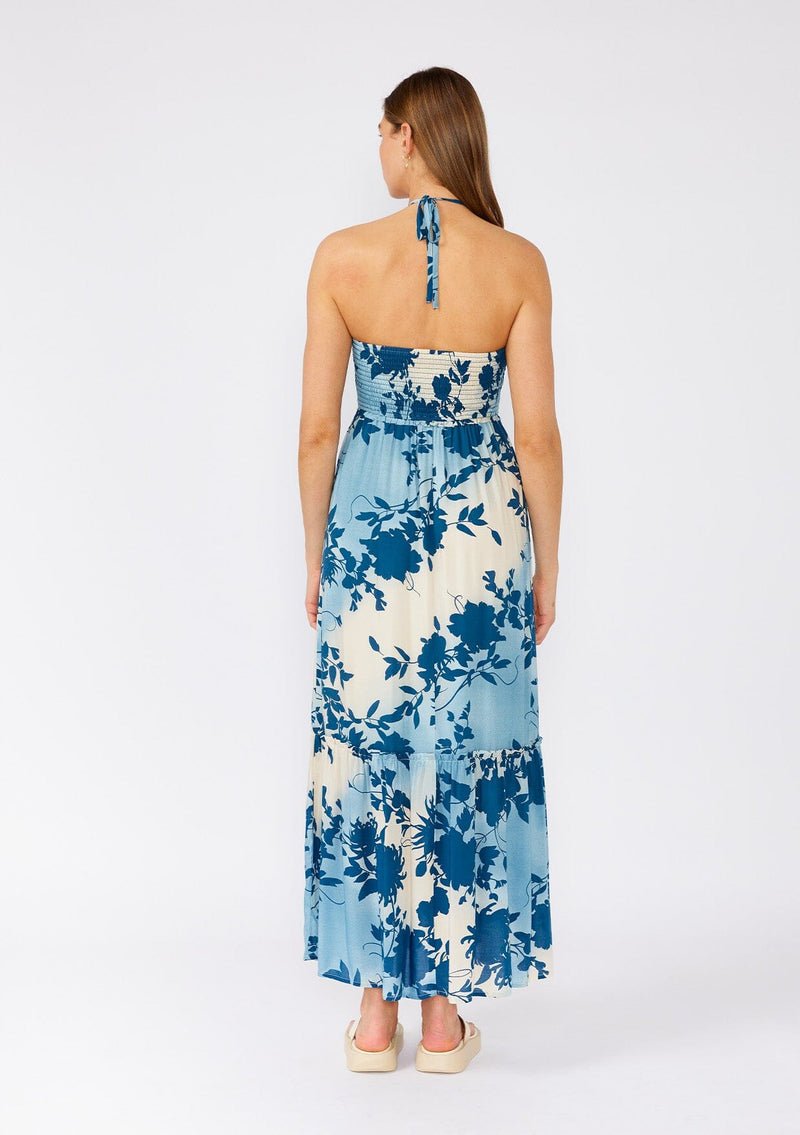 [Color: Navy/Dusty Blue] A back facing image of a brunette model wearing a dreamy bohemian halter maxi dress in a blue floral print. With a smocked elastic slim fit bodice, a long ruffles trimmed tiered skirt, and an adjustable halter tie neckline. 