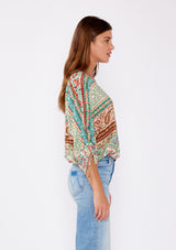 [Color: Jade/Natural] A brunette women wearing a multi print bohemian top with a v neckline, button front, ruched long sleeves, and elastic hemline. A teal and green causal summer top styled with denim for a summer to fall transition.