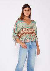 [Color: Jade/Natural] A brunette women wearing a multi print bohemian top with a v neckline, button front, ruched long sleeves, and elastic hemline. A teal and green causal summer top styled with denim for a summer to fall transition. 