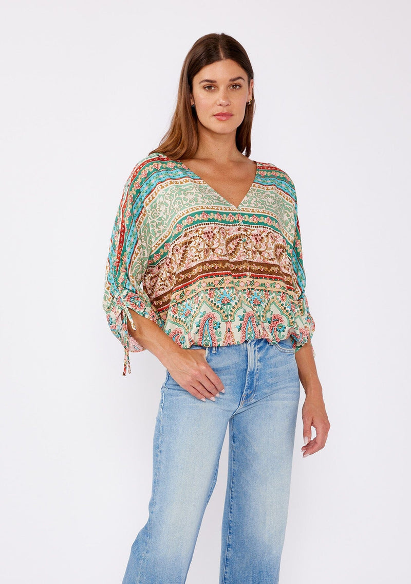 [Color: Jade/Natural] A brunette women wearing a multi print bohemian top with a v neckline, button front, ruched long sleeves, and elastic hemline. A teal and green causal summer top styled with denim for a summer to fall transition. 