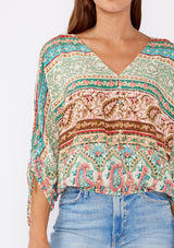 [Color: Jade/Natural] A brunette women wearing a multi print bohemian top with a v neckline, button front, ruched long sleeves, and elastic hemline. A teal and green causal summer top styled with denim for a summer to fall transition.