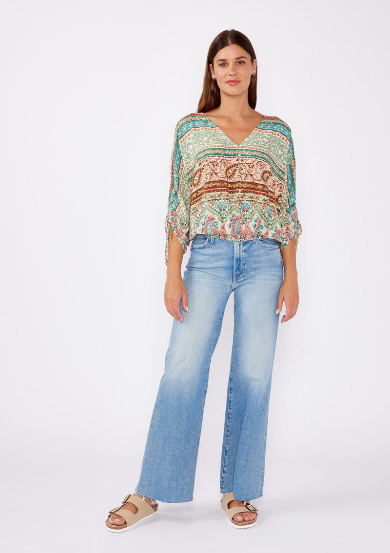 [Color: Jade/Natural] A brunette women wearing a multi print bohemian top with a v neckline, button front, ruched long sleeves, and elastic hemline. A teal and green causal summer top styled with denim for a summer to fall transition.