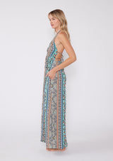 [Color: Natural/Tangerine] A side facing image of a blonde model wearing a sleeveless summer maxi dress in a bohemian mixed floral print, with gold metallic thread details. With a halter v neckline, an empire waist, and a strappy back with adjustable ties. 