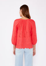 [Color: Coral] A brunette woman wearing an orange coral embroidered eyelet top with a split v neckline with ties, scalloped hem, and three quarter sleeve with button cuff. A see through top with no lining.