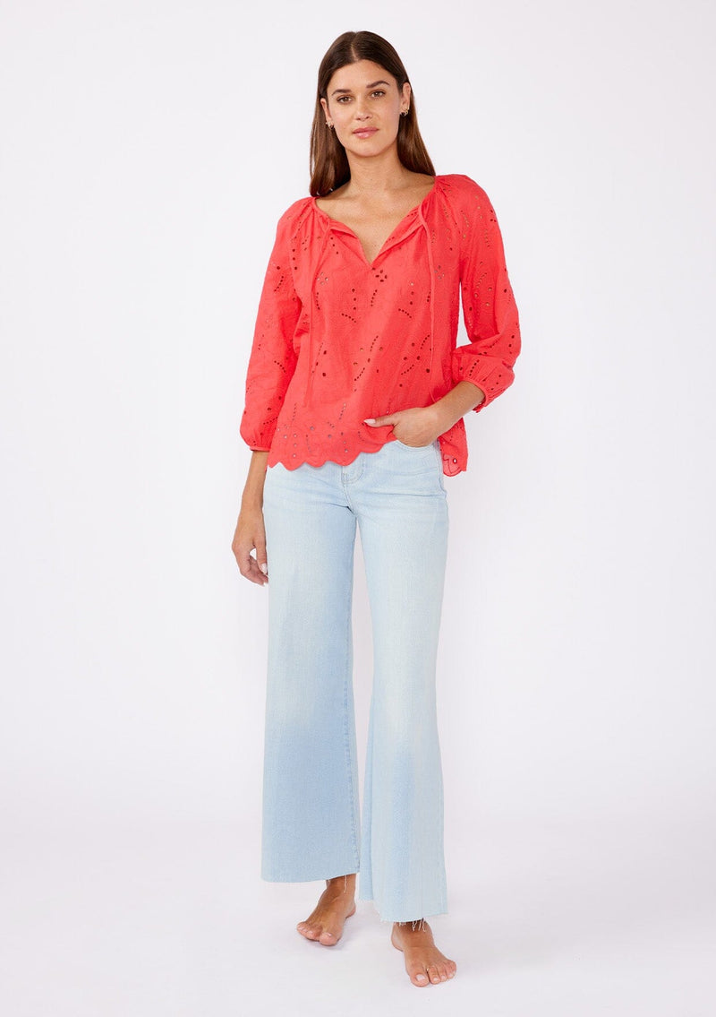 [Color: Coral] A brunette woman wearing an orange coral embroidered eyelet top with a split v neckline with ties, scalloped hem, and three quarter sleeve with button cuff. A see through top with no lining.