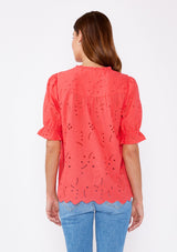 [Color: Coral] A brunette model wearing a coral orange top with a high round neckline, button front, and puff sleeves. Features a ruffled elastic cuff for added flair. A summer top with no lining for a comfortable breezy style for the summer.