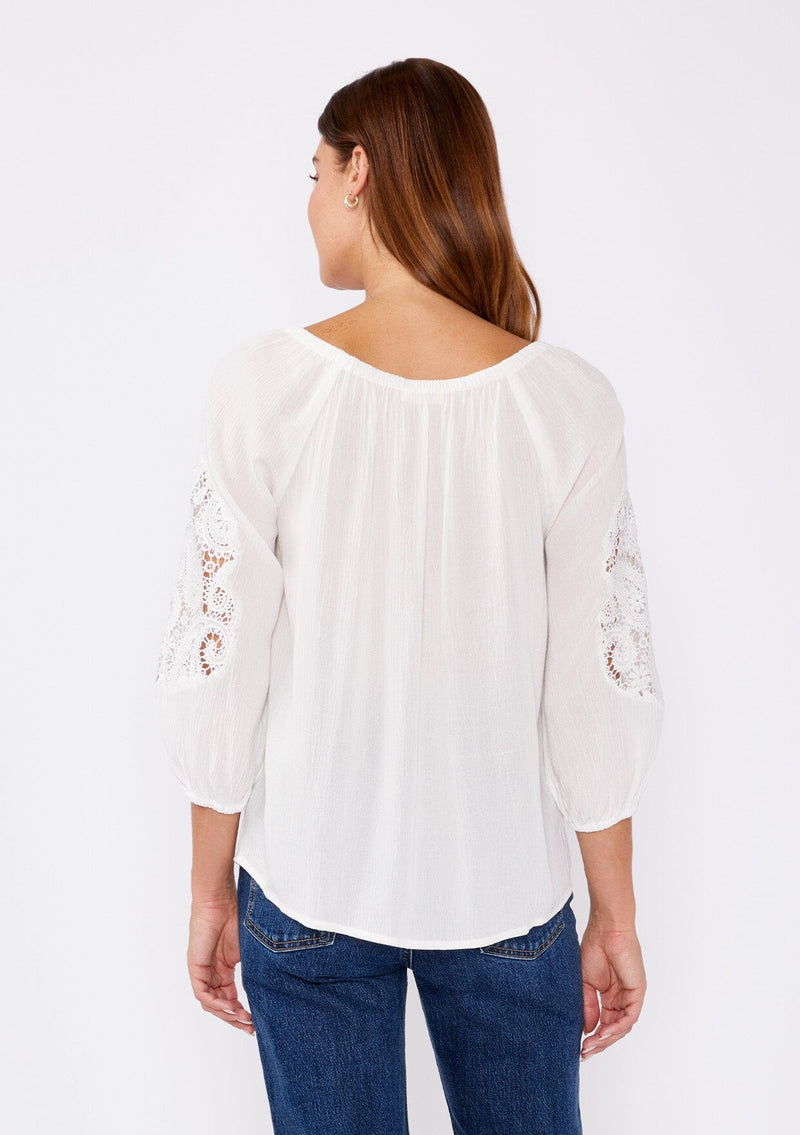 [Color: Off White] A brunette model wearing a casual white blouse with crochet lace detail. A summer top with an elastic neckline for on and off shoulder styling and three quarter length sleeves with elastic cuffs. A relaxed a flowy style perfect for casual outings.