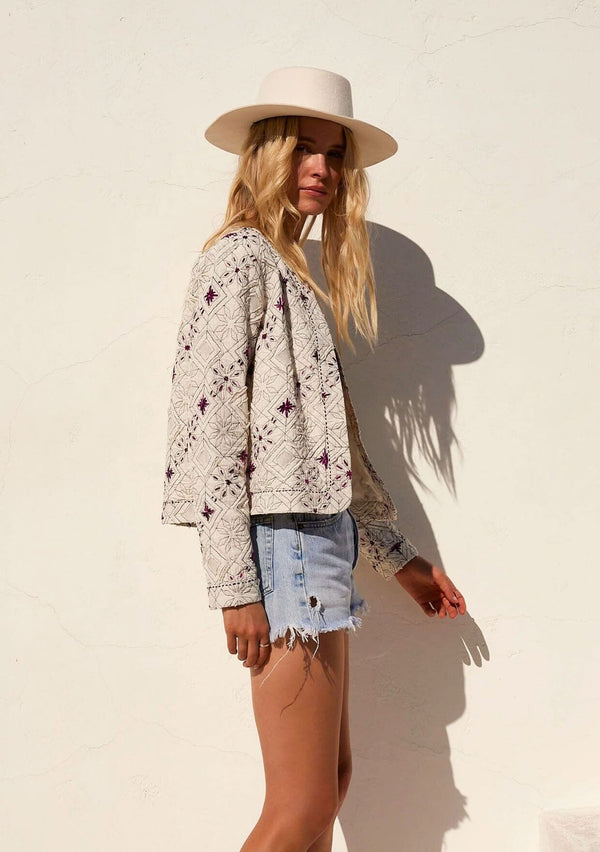 [Color: Natural/Fuchsia] A half body front facing image of a blonde model wearing a lightweight bohemian spring jacket in ivory with pink embroidered detail and contrast black thread detail. With long sleeves, an open front, and a cropped fit.