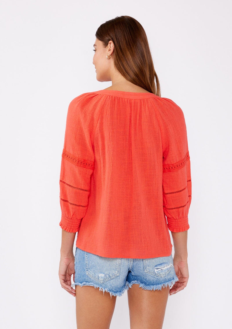 [Color: Coral] A brunette model wearing a textured cotton blouse for summer. Features a split v neckline, three quarter length sleeve, smocked cuff, and delicate lace trim inserts. A breathable and stylish cotton blouse for any occasion. 