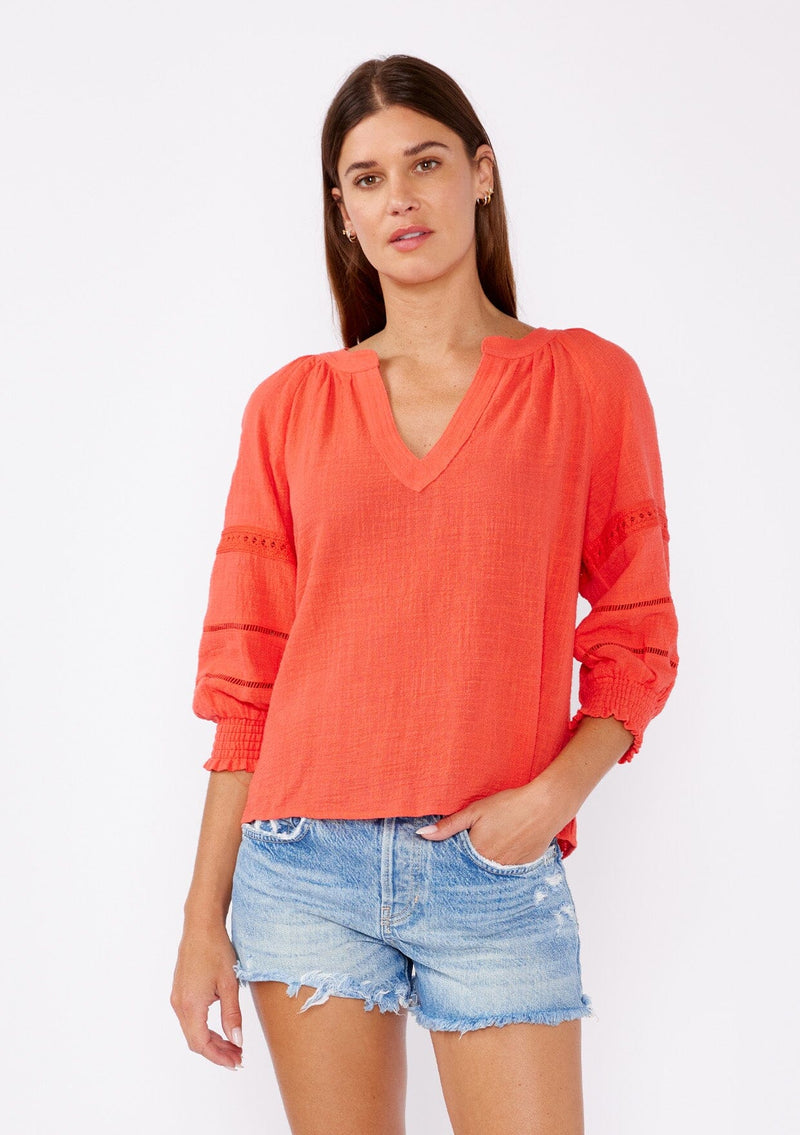 [Color: Coral] A brunette model wearing a textured cotton blouse for summer. Features a split v neckline, three quarter length sleeve, smocked cuff, and delicate lace trim inserts. A breathable and stylish cotton blouse for any occasion. 