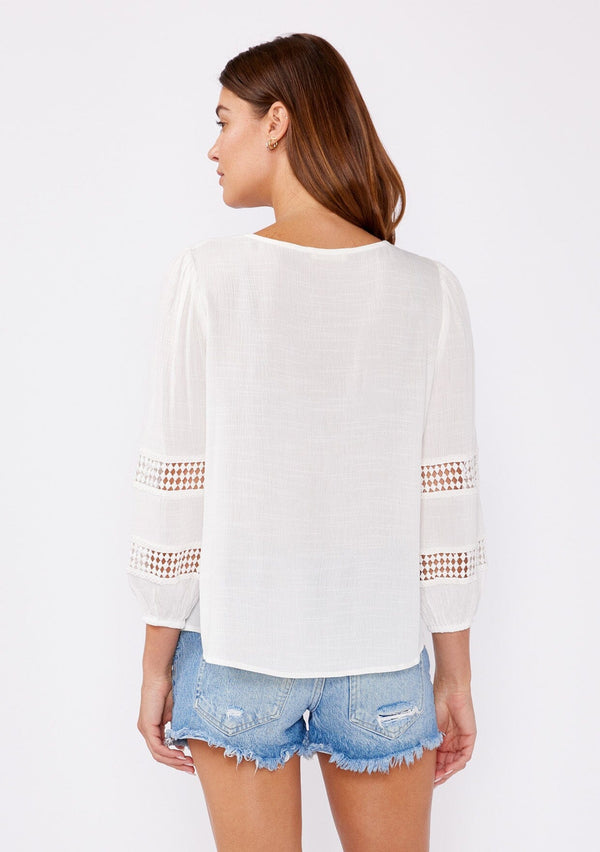 [Color: Natural] A brunette model wearing a white casual top with lace trim or crochet inserts. With a v neckline, long sleeve with elastic cuff, and a flowy fit designed with crinkled rayon fabric. A summer blouse perfectly styled with denim shorts.