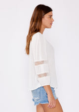 [Color: Natural] A brunette model wearing a white casual top with lace trim or crochet inserts. With a v neckline, long sleeve with elastic cuff, and a flowy fit designed with crinkled rayon fabric. A summer blouse perfectly styled with denim shorts.