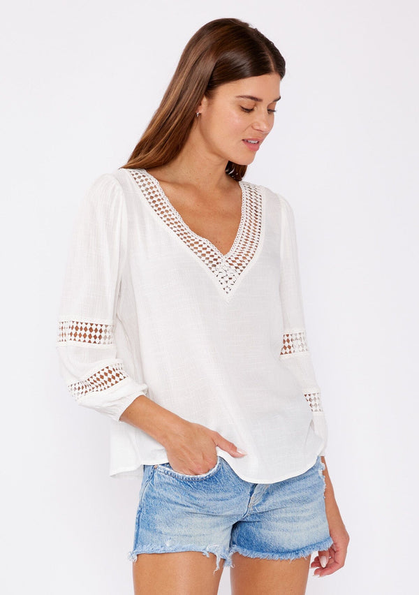 [Color: Natural] A brunette model wearing a white casual top with lace trim or crochet inserts. With a v neckline, long sleeve with elastic cuff, and a flowy fit designed with crinkled rayon fabric. A summer blouse perfectly styled with denim shorts. 