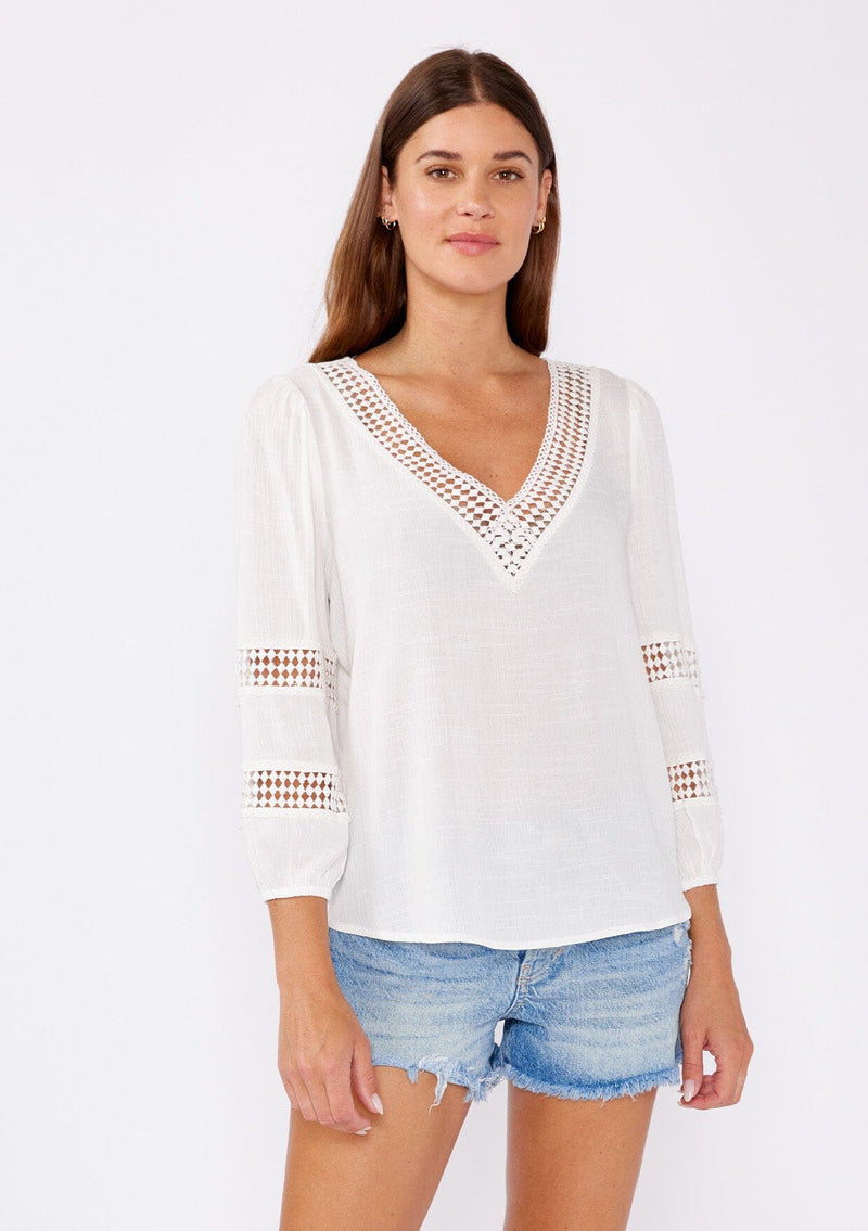 [Color: Natural] A brunette model wearing a white casual top with lace trim or crochet inserts. With a v neckline, long sleeve with elastic cuff, and a flowy fit designed with crinkled rayon fabric. A summer blouse perfectly styled with denim shorts.