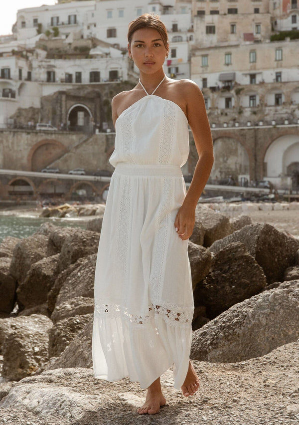[Color: Off White] A brunette model wearing a white ultra bohemian maxi dress for vacation. This summer maxi dress features a halter neckline and a comfortable smocked waist. This dress is also embellished with intricate lace details and embroidered eyelet. The perfect style for brunch or a tropical vacation.