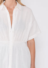[Color: White] A close up front facing image of a blonde model wearing a white tunic shirt with a collared neckline, a button front, short sleeves with a dropped shoulder, and a smocked elastic waist. 