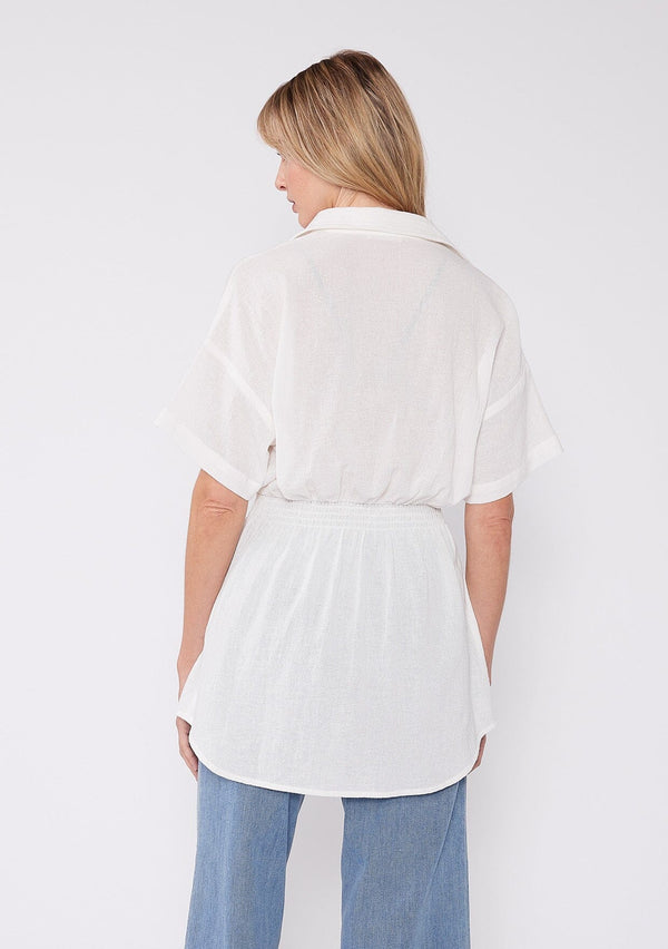 [Color: White] A back facing image of a blonde model wearing a white tunic shirt with a collared neckline, a button front, short sleeves with a dropped shoulder, and a smocked elastic waist. 
