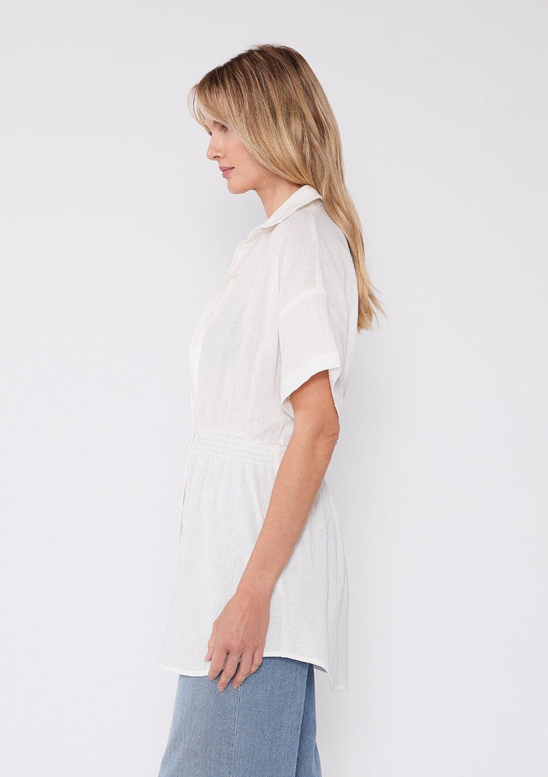[Color: White] A side facing image of a blonde model wearing a white tunic shirt with a collared neckline, a button front, short sleeves with a dropped shoulder, and a smocked elastic waist. 