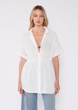 [Color: White] A half body front facing image of a blonde model wearing a white tunic shirt with a collared neckline, a button front, short sleeves with a dropped shoulder, and a smocked elastic waist. 