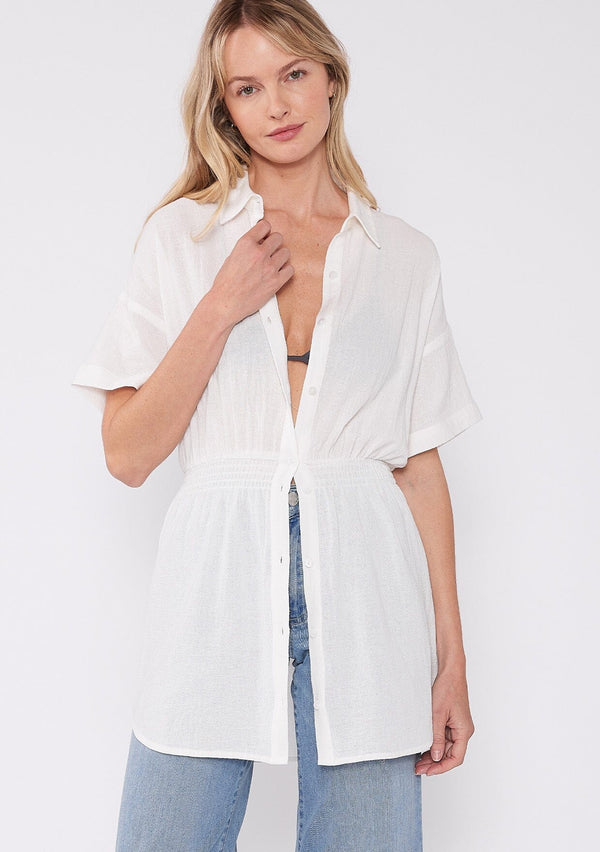 [Color: White] A front facing image of a blonde model wearing a white tunic shirt with a collared neckline, a button front, short sleeves with a dropped shoulder, and a smocked elastic waist. 