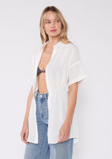 [Color: White] A half body side facing image of a blonde model wearing a white tunic shirt with a collared neckline, a button front, short sleeves with a dropped shoulder, and a smocked elastic waist. 
